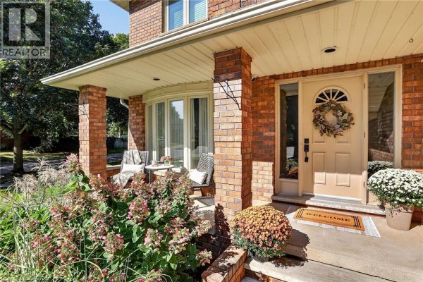 Covered front porch with brick pillars, featuring chairs, plants, and a welcome mat. - 64 Driftwood Court, Grimsby