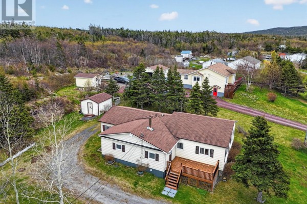 Aerial view of a beige bungalow with a wooden deck, surrounded by green grass and pine trees. - 64 Main Road, Broad Cove  Dildo