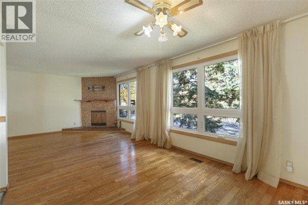Living room with a brick fireplace, hardwood floors, ceiling fan, and large windows with beige curtains. - 6418 7th Avenue N, Regina