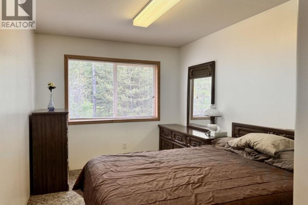 Bedroom with a dark wood dresser, a queen-size bed, and a window overlooking trees. - 6436 Erickson Road, 100 Mile House