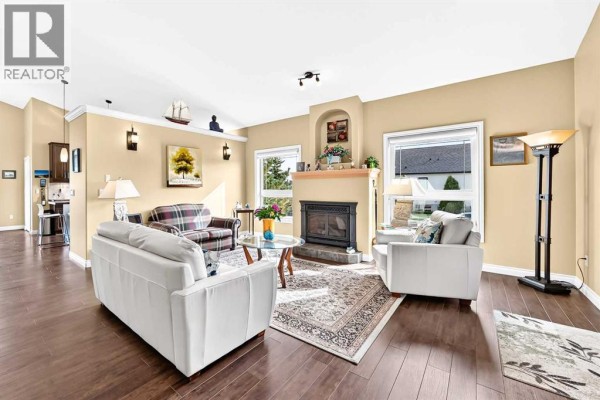 Living room with hardwood floors, a fireplace, two sofas, an armchair, and large windows with views of houses. - 6444 Cedar Way, Innisfail