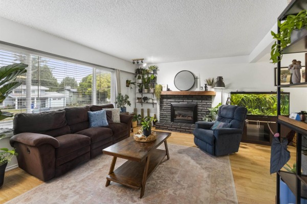 Living room featuring a dark brown sofa, recliner, fireplace, and large window. - 6486 Fern Street, Sardis South, Chilliwack