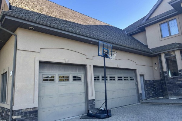 Side view of a beige house showing two grey garage doors with windows, a basketball hoop, and dark grey roof. - #65 53217 Rge Road 263, Rural Parkland County