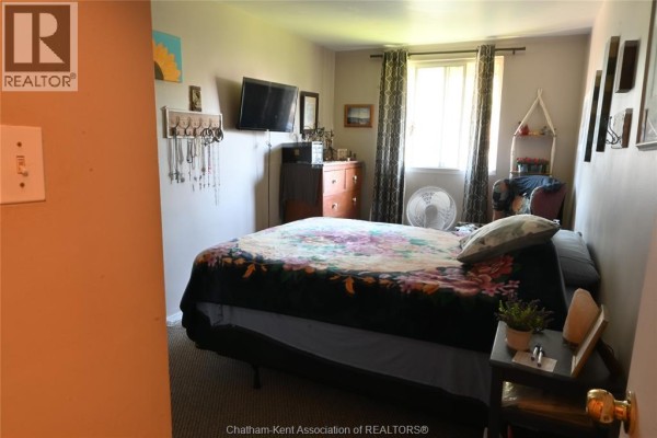 Bedroom with a queen-size bed, dresser, television, and window with curtains. - 65 Baldoon Road Unit# 11, Chatham