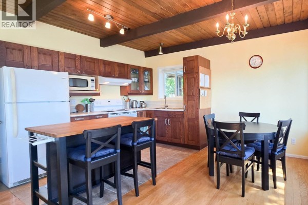 Kitchen with dark wood cabinets, white appliances, and an island with bar stools adjacent to a dining table with chairs. - 65 Esplanade Road, Keats Island