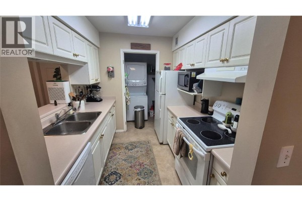 Kitchen with white cabinets, beige countertops, double sink, and a stacked washer and dryer visible. - 650 3 Street Sw Unit# 404, Salmon Arm