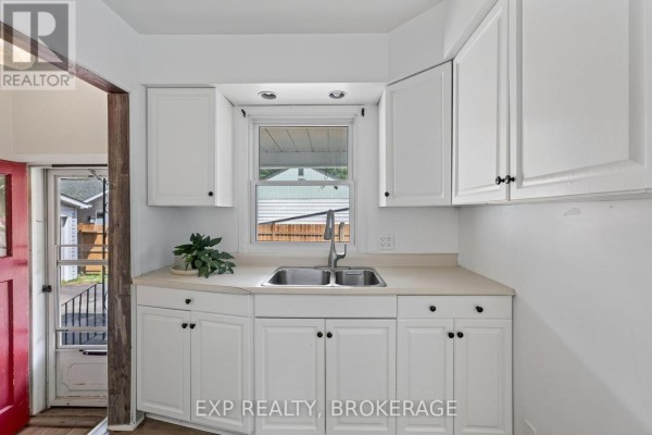 Close-up of white kitchen cabinets, double sink, and faucet with a window view. - 650 Hillside Drive, Gananoque
