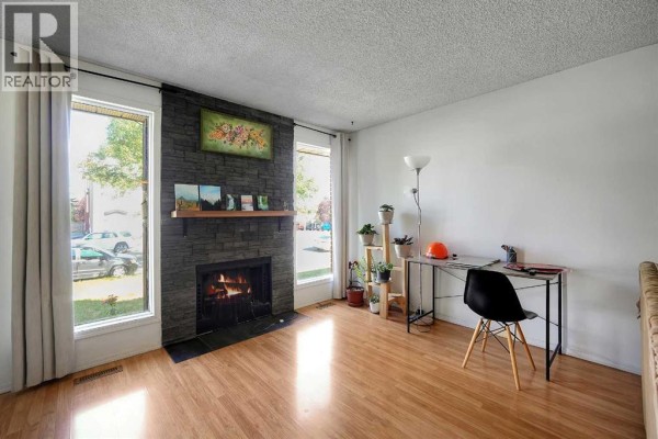 Living room featuring a stone fireplace, large windows, a desk, and light wood laminate flooring. - 6507 Rundlehorn Drive Ne, Calgary