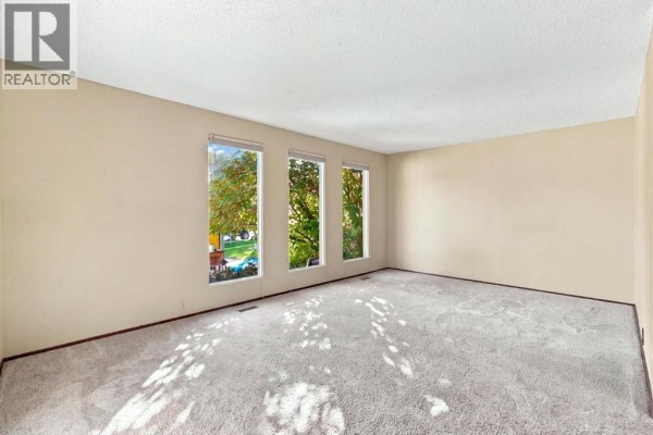 Empty living room with beige walls, plush carpet, and three large windows showing trees. - 652 Oakwood Place Sw, Calgary
