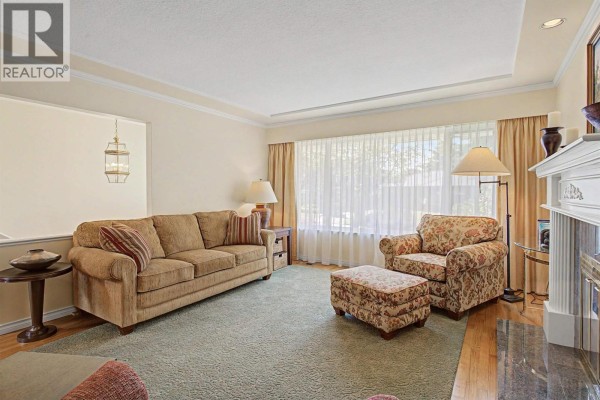Living room with a sofa, patterned armchair and ottoman, and a large window with sheer curtains. - 6541 Grant Street, Burnaby