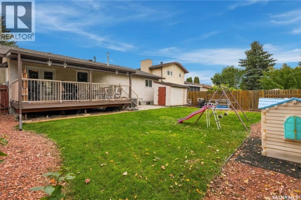 Backyard view with a wooden deck, grass, play structure, and a small toy house. - 6546 Rush Place, Regina
