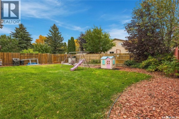 A fenced backyard with a green lawn, a play structure, and mature trees. - 6546 Rush Place, Regina