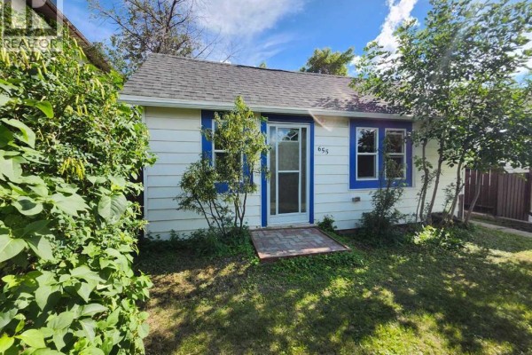Front view of a white bungalow with blue trim, a glass door, and a paved walkway. - 655 3 Avenue W, Drumheller