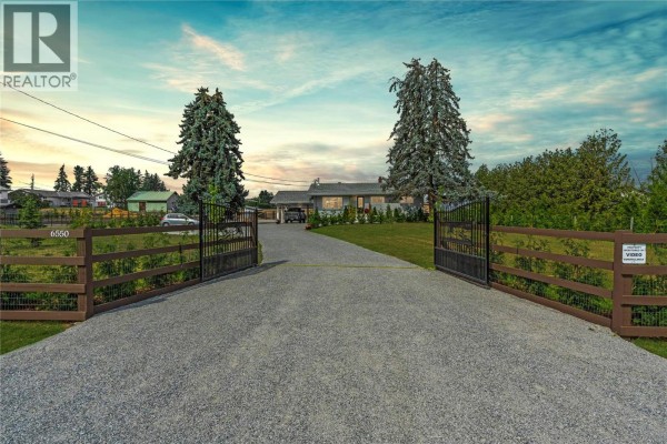 A long gravel driveway leads to a rancher home, framed by large gates and mature trees. - 6550 Herry Road, Vernon