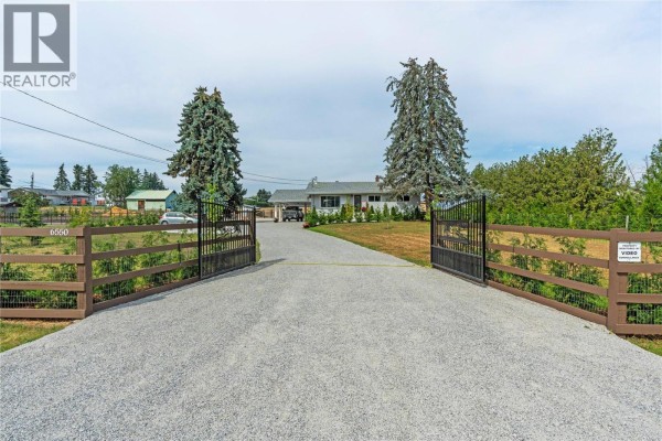 A gravel driveway leads through large gates to a rancher home with a manicured lawn and trees. - 6550 Herry Road, Vernon