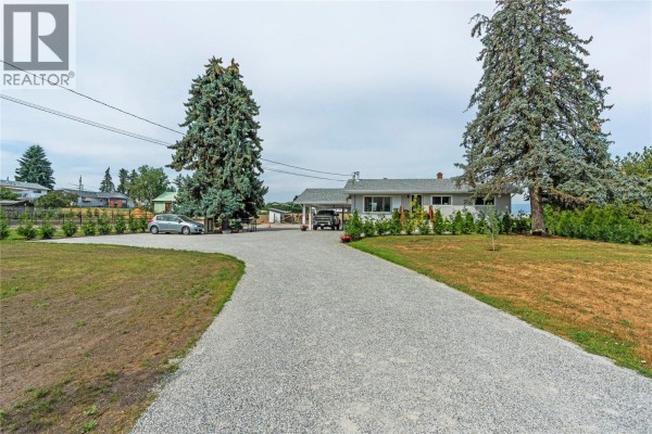 A gravel driveway curves towards a rancher home with a carport, flanked by green lawns and trees. - 6550 Herry Road, Vernon