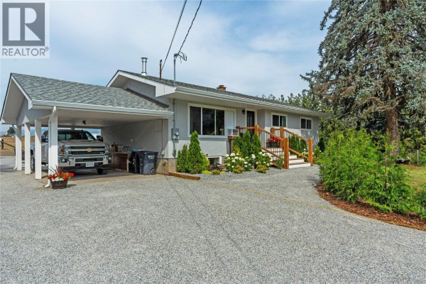 The front of a light grey rancher home with a carport, wooden deck, and manicured landscaping. - 6550 Herry Road, Vernon