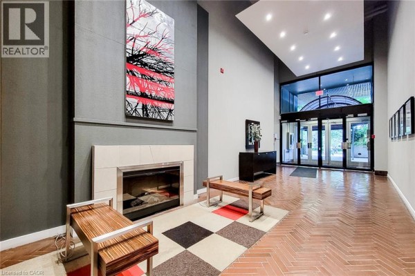 Building lobby with a modern fireplace, seating benches, abstract art, and glass entrance doors. - 66 Bay Street S Unit# 414, Hamilton