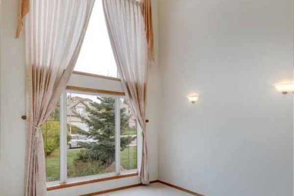 Large corner window with beige curtains overlooking a street and a green tree. - 662 Arbour Lake Drive Nw, Calgary