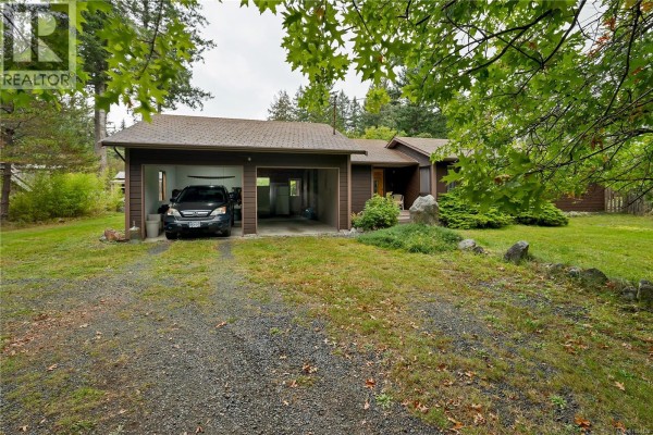 Front exterior of the house showing a dark brown siding, a gravel driveway, and a two-bay carport with a vehicle. - 665 Fox Rd, Quadra Island