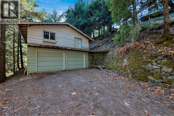 Detached garage with two light green doors and a dirt driveway on a wooded slope. - 6693 Genoa Bay Rd, Duncan