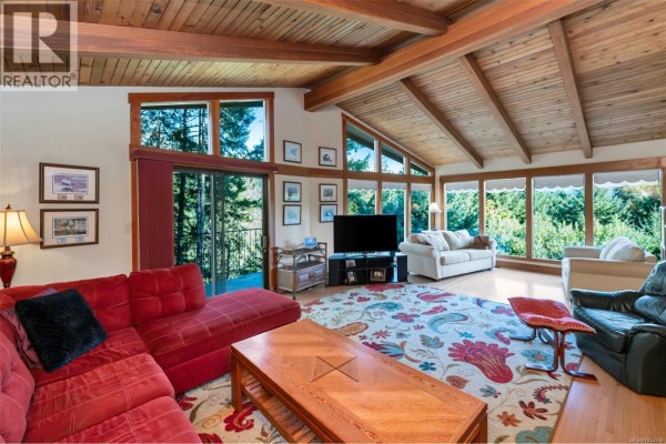 Living room with a red sectional, patterned rug, wood ceiling, and large windows overlooking trees. - 6693 Genoa Bay Rd, Duncan