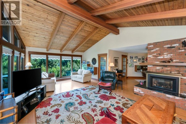 Living room with a brick fireplace, wood ceiling, and large windows facing a wooded area. - 6693 Genoa Bay Rd, Duncan