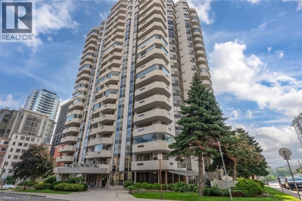 Tall beige condo building with balconies and evergreen trees - 67 Caroline Street S Unit# 13a Hamilton