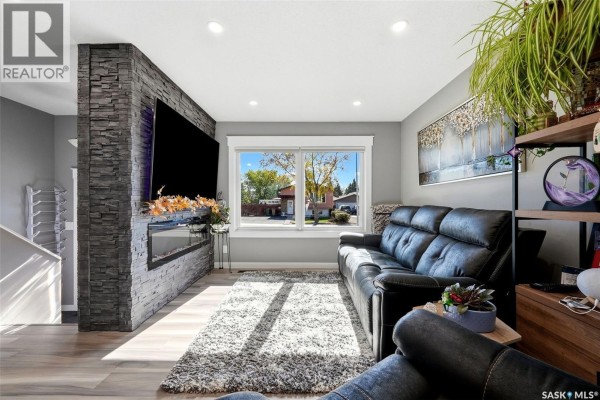 Living room with a large modern fireplace, television, black sofa, and large window overlooking the street. - 6702 Whelan Drive, Regina