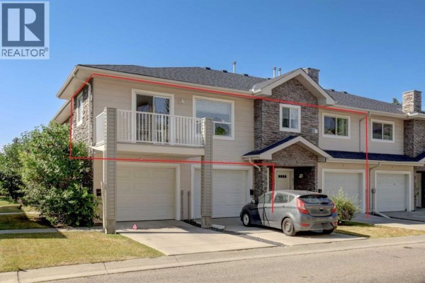 Exterior of a townhouse with beige siding, stone accents, and attached garages, showing a side view with a balcony. - 6711 Pinecliff Grove Ne, Calgary