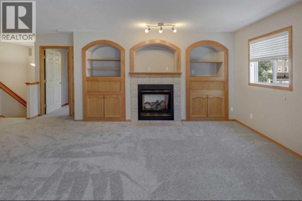 Living room with grey carpet, fireplace, and built-in oak cabinets and shelving, with a window view. - 6711 Pinecliff Grove Ne, Calgary