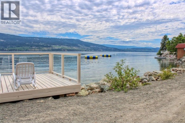 Waterfront view with a wooden deck, lounge chair, and distant mountains under a cloudy sky. - 6742 Marbella Loop Unit# 302, Kelowna
