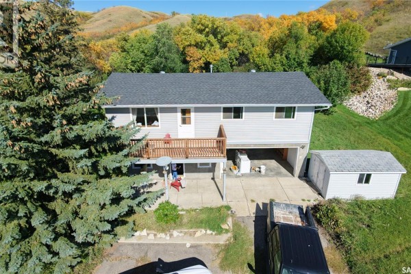 Exterior of a grey house with a wooden deck, attached garage, and a small shed against a backdrop of rolling hills. - 675 Fort San Road, Fort San