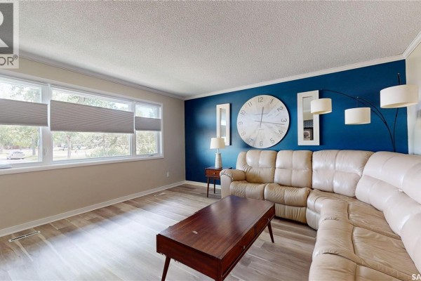 Living room with a sectional sofa, dark blue accent wall with a large clock, and light wood-look flooring. - 68 Cameron Crescent, Regina