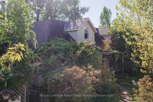 Side exterior of a house with a steep roof, dormer, and overgrown plants on a sunny day. - 68 Victoria Street, Ashfield Colborne Wawanosh