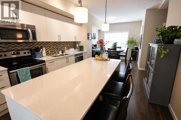 Living room with a grey sectional sofa, large rug, and sliding glass doors to a backyard. - 680 Old Meadows Road Unit# 85, Kelowna