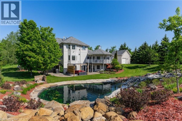 Backyard with large pond, rock landscaping, trees, and distant outbuilding. - 6805 Concession Rd 2, Puslinch