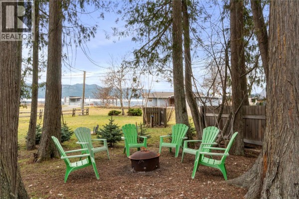 Green Adirondack chairs and fire pit on a gravel area with tall trees and a lake view. - 6807 Squilax - Anglemont Road, Magna Bay