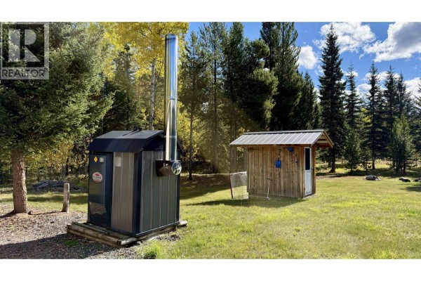Outdoor wood boiler with a metal chimney next to a small wooden shed. - 6836 Fawn Creek Road, Lone Butte