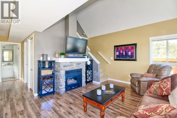 Living room with gas fireplace, television, sofa, armchair, and wood-look flooring. - 6871 Santiago Loop Unit# 115, Kelowna