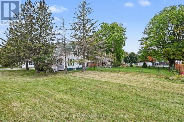 View of the house from the side yard, showing the white exterior, green shutters, and chain-link fence. - 69 Main Street, North Dundas