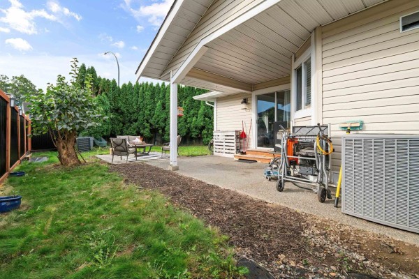 Covered concrete patio with outdoor seating, adjacent to a grass lawn and tall green hedges. - 6900 Oakwood Drive, Agassiz, Agassiz