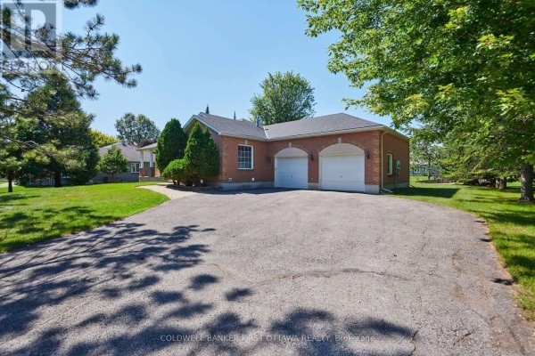 Rear view of the red brick bungalow showing a paved driveway and a three-car garage. - 6983 Lakes Park Drive, Ottawa