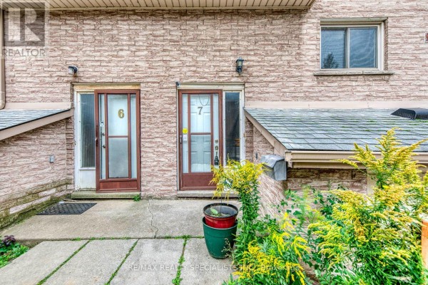 Front entrance of townhouse unit 7, showing a doorway, concrete walkway, and surrounding greenery. - 7 - 325 Mary Street, Hamilton