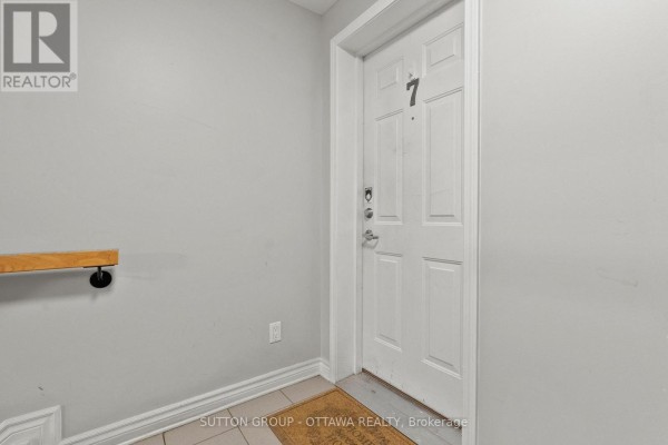 Interior view of a white apartment door labeled '7' with a doormat and light grey walls. - 7 - 565 Stonefield Private, Ottawa