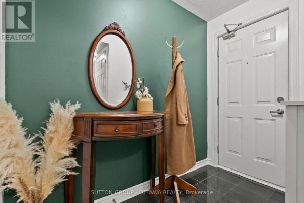 Entrance hallway with dark green walls, a wooden console table, oval mirror, coat rack with coat, and a white door. - 7 - 565 Stonefield Private, Ottawa