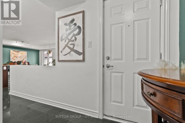 View of an apartment entryway with a white door, artwork on white wall, and glimpse into living area. - 7 - 565 Stonefield Private, Ottawa