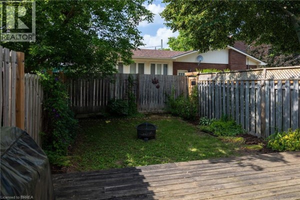 Fenced backyard with wooden deck, green lawn, and a fire pit surrounded by trees. - 7 Graystone Court, Paris