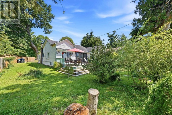 View of a white cottage from the side, showing the deck, lawn, and surrounding trees. - 7 Oliver Drive, Tiny