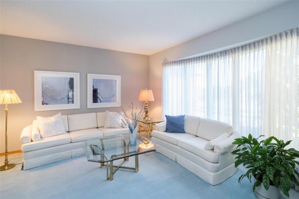 Living room with two cream sofas, glass coffee table, blue carpet, and floor-to-ceiling windows. - 7 Princemere Road, Winnipeg